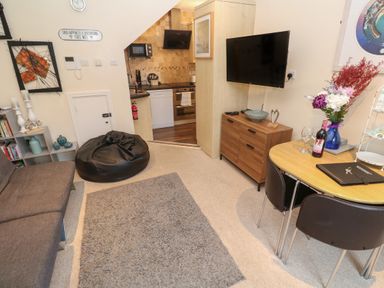 A living room with sofa bean bag table chairs flowers and TV and view into kitchen at 5 Lyonesse Apartments in St Ives