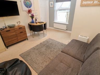 A living room with a gray sofa a wooden cabinet a small dining table with chairs and a wall-mounted television at 5 Lyonesse Apartments in St Ives