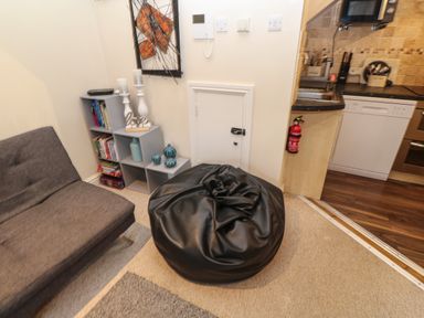A small living area with a gray sofa black bean bag bookshelf with decorations and a fire extinguisher near the kitchen at 5 Lyonesse Apartments in St Ives