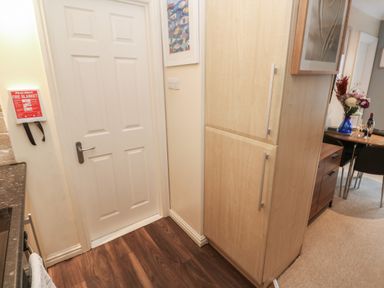 A kitchen corner with a white door a wooden cabinet a fire blanket on the wall and a dining area with a vase of flowers at 5 Lyonesse Apartments in St Ives