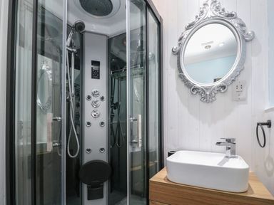 A bathroom with a glass shower enclosure a rectangular sink on a wooden vanity and an ornate round mirror at 5 Lyonesse Apartments in St Ives