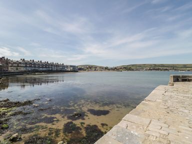A coastal scene with a stone pier and calm water near a row of buildings and hills in the distance at Old Harry Cottage 16 in Swanage
