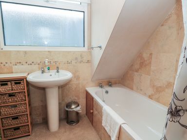 A bathroom with a pedestal sink a wicker drawer unit a metal trash can a bathtub with a towel and a curtain at Old Harry Cottage 16 in Swanage