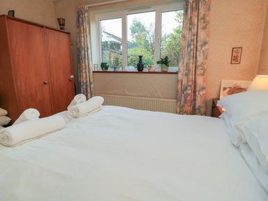 A bedroom with a bed made with white linens rolled towels on the bed a wooden wardrobe against the wall and a window with curtains at Old Harry Cottage 16 Swanage