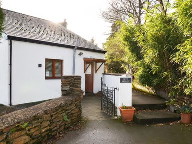 A small white cottage with a brown door and window behind a stone wall and metal gate surrounded by greenery at Artists Cottage in Minffordd near Penrhyndeudraeth