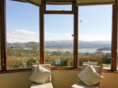 A room with two chairs having cushions and a vase with flowers on the windowsill overlooking trees and mountains at Artists Cottage in Minffordd near Penrhyndeudraeth