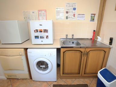 A laundry room with a washing machine, dryer, two mini fridges, a sink with cabinets, a blue trash bin, and information posters on the wall at Artists Cottage in Minffordd near Penrhyndeudraeth