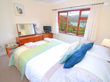A bedroom with a bed with pillows beside a wooden dresser with a mirror and a window showing trees and hills at Artists Cottage in Minffordd near Penrhyndeudraeth