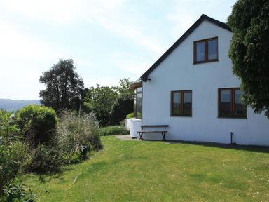 A white house with brown window frames and a bench outside on a green lawn with trees and bushes at Artists Cottage in Minffordd near Penrhyndeudraeth