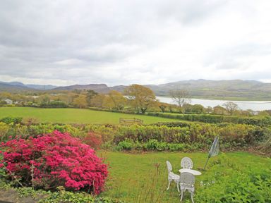 A garden with a white metal table and chairs surrounded by green grass and bushes overlooking fields trees water and hills at Artists Cottage in Minffordd near Penrhyndeudraeth