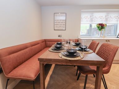 A dining area with a wooden table set with black dishes and glassware plus brown cushioned bench and chairs near a window with floral curtains at Beudy Pwll Pridd in Llangollen
