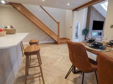 A dining area with brown chairs and a table set with black dishes next to a kitchen counter with wooden stools and a staircase in the background at Beudy Pwll Pridd in Llangollen