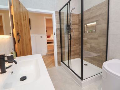 A bathroom with a sink and faucet a glass shower enclosure with wooden wall panels and a wooden door open to an adjacent room at Beudy Pwll Pridd in Llangollen