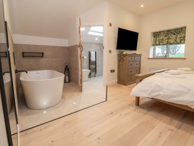 A bedroom with a wooden bed a chest of drawers and a wall-mounted TV next to an open door leading to a bathroom with a freestanding bathtub and a toilet at Beudy Pwll Pridd in Llangollen