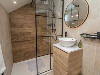 A bathroom with a walk-in shower with wood wall panels a circular mirror above a wooden vanity with a white basin at Beudy Pwll Pridd in Llangollen