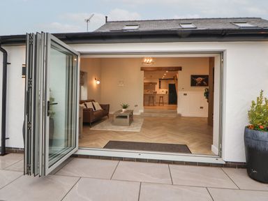 An open folding glass door leading to a living room with a brown couch a coffee table and a kitchen area in the background at Beudy Pwll Pridd in Llangollen