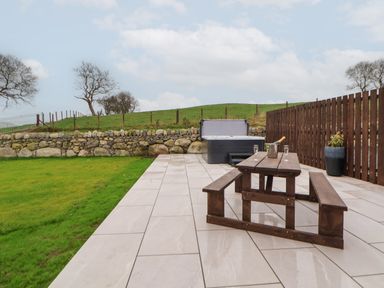 A patio with a wooden picnic table and bench with a bottle and glasses and a hot tub near a stone wall at Beudy Pwll Pridd in Llangollen