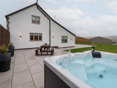 An outdoor patio with a hot tub holding two glasses and a bottle and a wooden picnic table near a white house at Beudy Pwll Pridd in Llangollen