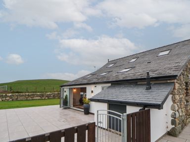 A stone and white house with multiple skylights and an open glass door next to a tiled patio with a metal fence and green hills in the background at Beudy Pwll Pridd in Llangollen