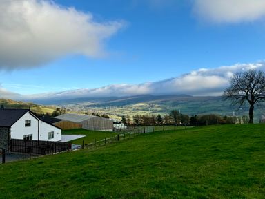 A white house with a fenced yard next to a large barn and green fields with trees and hills in the background at Beudy Pwll Pridd in Llangollen