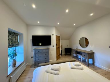 A bedroom with a bed, folded towels, a chest of drawers with a wall-mounted TV, a window with a potted plant, a dressing table with a round mirror, and a wooden door at Beudy Pwll Pridd in Llangollen