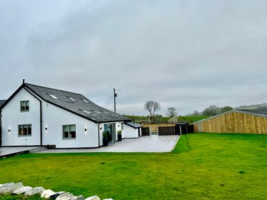 A white house with a slate roof and multiple windows next to a tiled patio and green lawn at Beudy Pwll Pridd in Llangollen