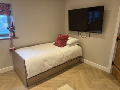 A single bed with white bedding and a red cushion next to a window with a stuffed reindeer toy on the sill and a wall-mounted television in a room at Beudy Pwll Pridd in Llangollen