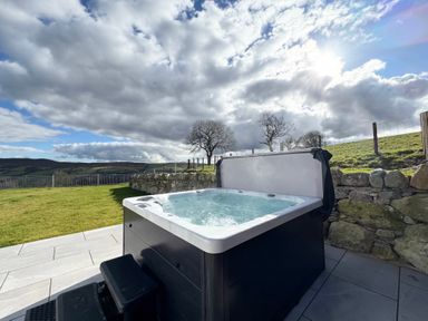 A hot tub on a paved patio next to a stone wall with grass and hills in the background at Beudy Pwll Pridd in Llangollen