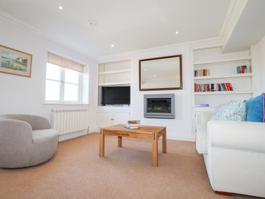 A living room with a wooden coffee table books a gray armchair a window a wall-mounted fireplace a mirror built-in shelves a television and a white sofa with cushions at 6 Willows Green in Aldeburgh