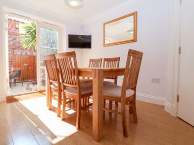A dining room with a wooden table and six chairs near glass doors with a wall-mounted TV and mirror at 6 Willows Green in Aldeburgh