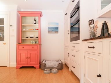 A kitchen corner with a coral cabinet holding dishes a pet bed with two bowls and white built-in cabinets with an oven at 6 Willows Green Aldeburgh