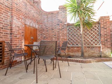An outdoor patio with a metal table and chairs a brick wall a small tree and a wooden door at 6 Willows Green in Aldeburgh
