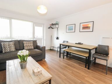 A living area with a dark sofa with patterned cushions a wooden coffee table with flowers a wooden dining table a bench two chairs and wall art at 102 Waterside Park in Corton