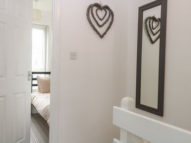 A hallway with a wall mirror and heart-shaped wall decoration next to a door opening to a bedroom with a bed and window at 102 Waterside Park in Corton