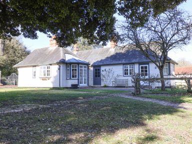 An exterior view of a single-story house with white wooden siding and multiple chimneys surrounded by grass and trees at Holm Oak Lodge in Tendring near Weeley