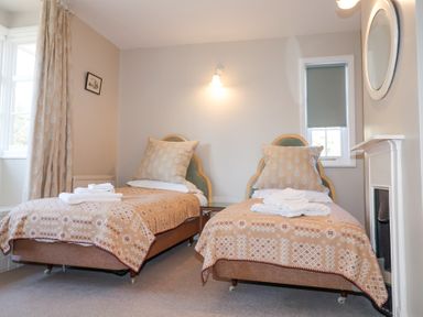 A bedroom with two single beds with patterned blankets and pillows a window with curtains and a window with a roller blind a wall light and a round mirror above a fireplace at Holm Oak Lodge in Tendring near Weeley