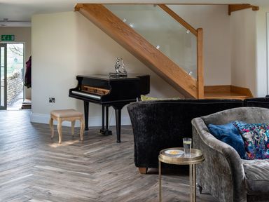 A black piano with a stool under a wooden staircase next to a gray sofa with blue and patterned cushions and a small round table with a glass and bowl at High View Barn in Sedbergh