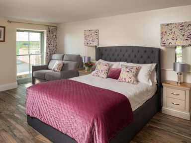 A bedroom with a bed covered in a purple blanket and pillows a gray armchair next to a window and two nightstands with lamps at High View Barn in Sedbergh