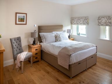 A bedroom with a double bed two bedside tables with lamps a gray cushioned chair and two windows with patterned blinds at High View Barn in Sedbergh
