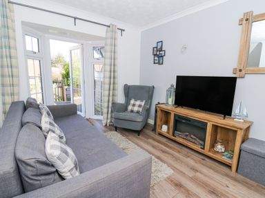 A living room with a gray sofa and armchair a wooden media unit with a TV and a bay window with doors and curtains at The Cwtch in Aberporth