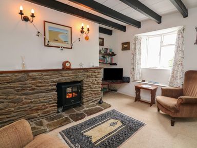 A living room with a stone fireplace a small stove an armchair a wooden table a patterned rug and a window with floral curtains at Vine Cottage in East Prawle