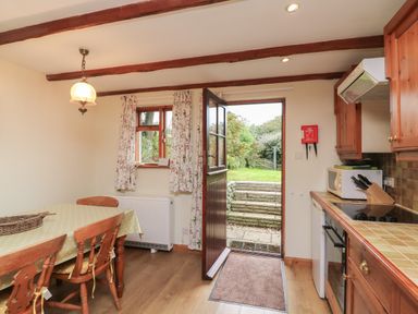 A kitchen with wooden cabinets a dining table with chairs and an open door leading to a garden at Vine Cottage in East Prawle