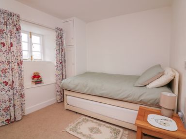 A small bedroom with a single bed green bedding floral curtains a window a wooden nightstand with a lamp and a small rug at Vine Cottage in East Prawle