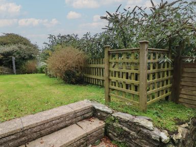 A garden with stone steps leading to a lawn and a wooden fence with bushes at Vine Cottage in East Prawle