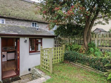 A backyard garden area with a small fenced section a tree and a view into a kitchen at Vine Cottage in East Prawle
