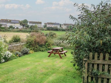 A garden with a wooden picnic table and a fence with houses in the background at Vine Cottage in East Prawle
