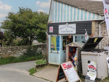 Small shop with sign Piglet Stores and Emporium outdoor menus and storage shed next to a stone wall and tree at Vine Cottage in East Prawle