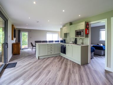 A kitchen with light green cabinets and an island open to a dining area with chairs and a small bedroom visible at Orchard Lodge in Maerdy near Corwen