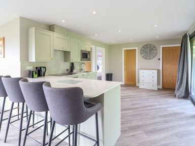 A kitchen with a breakfast bar and four grey bar stools two wooden doors a small white chest of drawers and a large clock on the wall at Orchard Lodge in Maerdy near Corwen