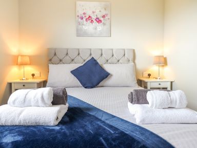 A bedroom with a bed with blue and white bedding and folded towels on it with two bedside tables with lamps and a floral painting on the wall at Orchard Lodge in Maerdy near Corwen